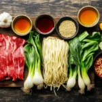 A top-down view of fresh ingredients for making sticky beef noodles, beautifully arranged on a rustic wooden surface. Thinly sliced raw flank steak marinated in soy sauce and sesame oil, fresh egg noodles, chopped garlic and ginger, green onions, bell peppers, and bok choy. Small bowls filled with soy sauce, hoisin sauce, oyster sauce, honey, and cornstarch